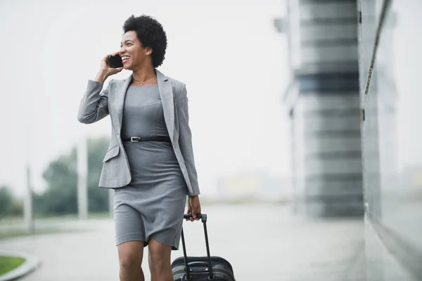 A cheerful black business woman walking with her luggage and talking on her smart phone outside of the airport.