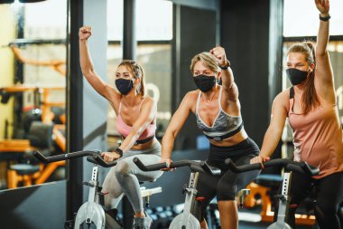 Koruyucu maskeli üç formda bayan arkadaştan oluşan bir grup Covid-19 salgını sırasında spor bisikleti eğitimi alıyorlar..