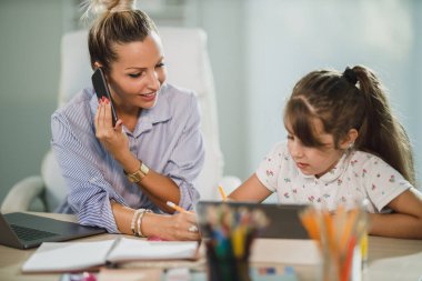 Güzel, meşgul, bekar bir anne evde akıllı telefonuyla konuşurken tatlı kızına ödevlerinde yardım ediyor..