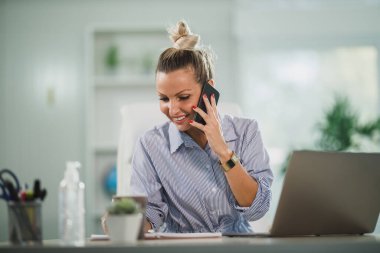 Ofisinde oturan ve bilgisayarında çalışırken cep telefonuyla konuşan çekici bir iş kadını..