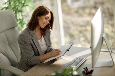 Çekici bir iş kadını, ofisindeki bilgisayarı kullanırken bir not defterine bir şeyler yazıyor..