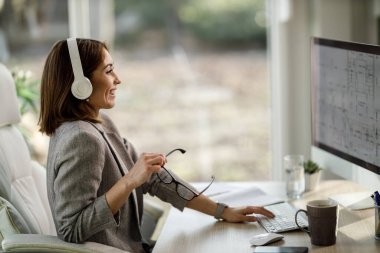 Neşeli bir iş kadını evinde kulaklıklarıyla tek başına oturuyor ve bilgisayarla çalışıyor..