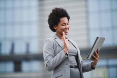 Bir şirket binasının önünde dijital tabletle konuşan gülümseyen zenci bir iş kadını portresi..