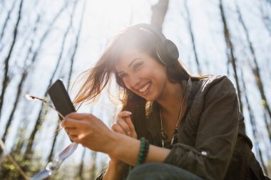 Doğada müzik dinlerken akıllı telefonunu kullanan gülümseyen genç bir kadının fotoğrafı..