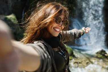 Özensiz genç bir kadın güzel şelalenin manzarasının tadını çıkarırken selfie çekiyor..