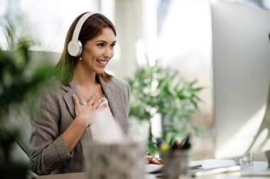 Kulaklıklı çekici bir kadın ofisinde tek başına oturuyor ve internet konferansı için bilgisayar kullanıyor..