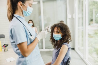 Hemşire elinde şırınga tutuyor ve maske takan genç bir kadının omzuna Covid-19 aşısı yapıyor..