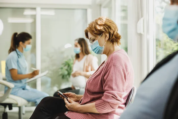 Senior woman with a face mask sitting in a waiting room of a hospital and surfing at her smartphone.