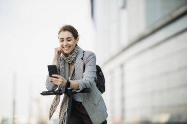 Şehirde elektrikli scooterla gezerken akıllı telefon kullanan genç bir iş kadını..