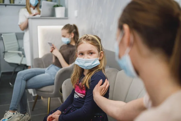 Pensive little girl and her mom with face mask in waiting room at dentist's office.