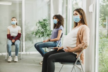 Yüzü maskeli genç hamile bir kadın bekleme odasında oturmuş jinekolog muayenesini bekliyor..