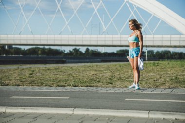 Orta yaşlı fit bir kadın nehir köprüsünün yanında sıkı bir eğitimden sonra dinleniyor ve müzik dinliyor..