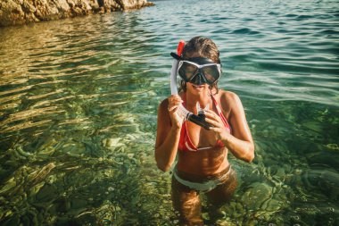 Dalgıç maskesi ve şnorkeliyle çekici bir kadın sahilde eğleniyor ve şnorkelle yüzmeye hazırlanıyor..