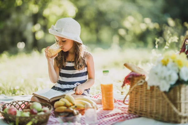 一个可爱的微笑的小女孩 一边喝着橙汁 一边看书 一边享受野外野餐 — 图库照片