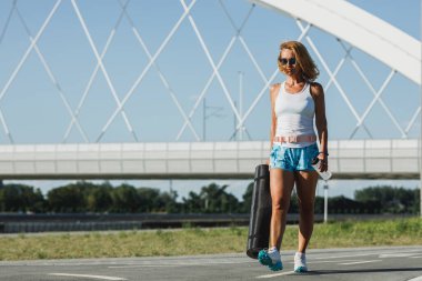 Orta yaşlı, sağlıklı bir kadın, yoga minderi tutuyor ve sıkı çalışmadan önce şehirde nehrin kenarında yürüyor..