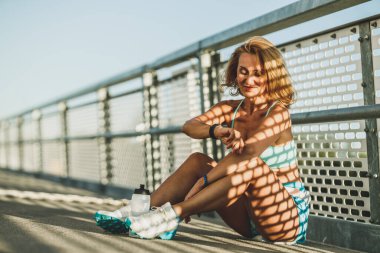 Orta yaşlı sporcu kadın köprüdeki eğitimden sonra dinlenirken akıllı saatlere bakıyor..