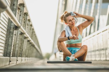 Orta yaşlı sporcu kadın köprüde çalıştıktan sonra dinlenirken akıllı saate bakıyor..