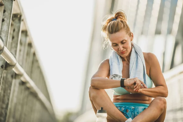 上了年纪的女运动员在桥上进行室外训练后 一边在外面休息 一边看着聪明的手表 — 图库照片