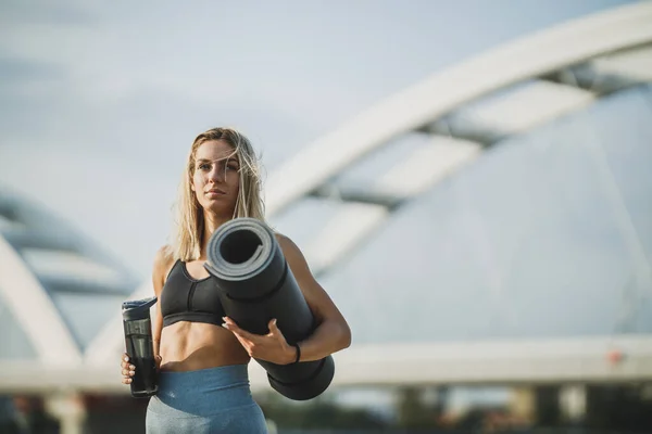 Portrait of a beautiful fit woman in sports wear holding a yoga mat and a bottle of water and walking near to river, in the city before training outdoor.