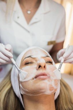 Güzellik uzmanı maske takıyor ve güzellik salonunda titizlikle yüz bakımı yapıyor.. 