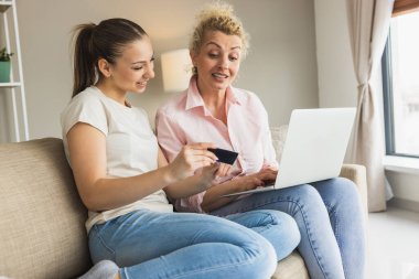 Neşeli anne ve kızı online alışveriş için bir dizüstü bilgisayar kullanarak birlikte oturuyorlar..
