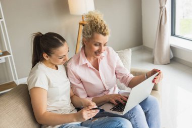Neşeli anne ve kızı online alışveriş için bir dizüstü bilgisayar kullanarak birlikte oturuyorlar..