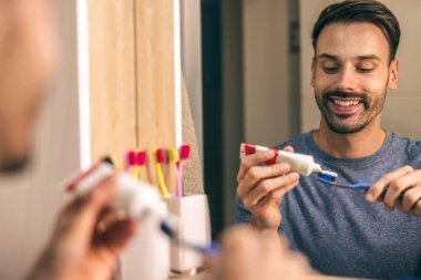 Bir adam dişlerini banyo aynasının önünde fırçalar. Temizlik alışkanlıklarını ve sabah rutinini yansıtır..