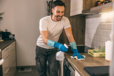 Adam mutfak tezgahını bez ve sprey şişeyle silip dezenfekte ediyor..