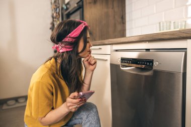 Evdeki mutfağında bulaşık makinesini kontrol etmek ve açmak için akıllı telefon kullanan güzel bir kadın..