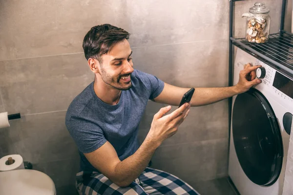 Genç adam evdeki banyoda çamaşır makinesini kontrol etmek için telefonunu kullanıyor..