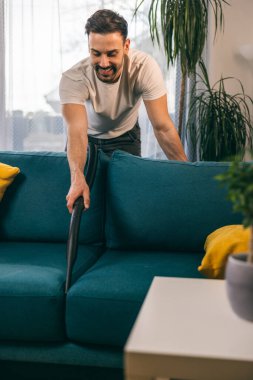 Genç adam oturma odasında kanepesini temizliyor ve süpürüyor..