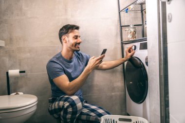 Genç adam evdeki banyoda çamaşır makinesini kontrol etmek için telefonunu kullanıyor..