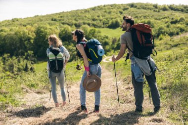 Güneşli bir günde sırt çantaları ve yürüyüş direkleriyle yürüyüş yapan üç mutlu arkadaş..
