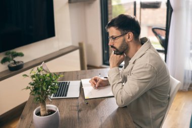 Gözlüklü genç adam ev ofisinde dizüstü bilgisayarla çalışıyor..