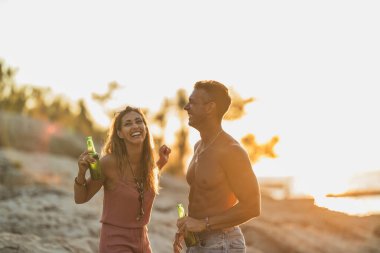 Young couple drinking beer on the beach by the sea. Enjoying in beautiful sunset and their love.