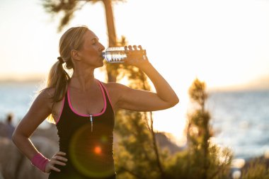 Formda bir orta yaşlı kadın koşucu deniz kıyısında koştuktan sonra su içiyor..