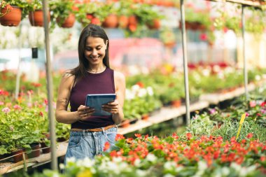 A smiling woman engaging with a tablet device surrounded by vibrant flowers in a greenhouse.