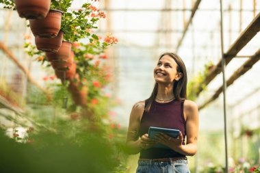 Gülümseyen bir kadın bir tabletle meşgul. Serada canlı çiçeklerle çevrili..