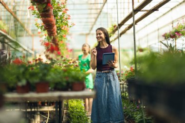 A smiling woman engaging with a tablet device surrounded by vibrant flowers in a greenhouse.
