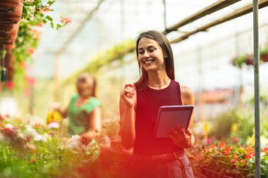 A smiling woman engaging with a tablet device surrounded by vibrant flowers in a greenhouse.