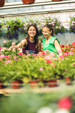 İki neşeli kadın güneşli bir seranın içinde çiçeklerle dolu canlı bir bahçenin keyfini çıkarıyorlar...