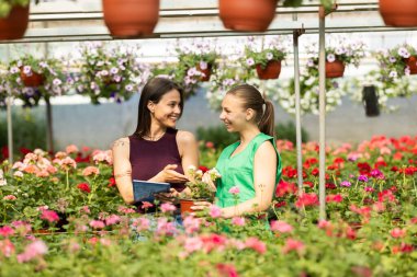 İki neşeli kadın güneşli bir seranın içinde çiçeklerle dolu canlı bir bahçenin keyfini çıkarıyorlar...