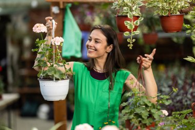 Mutlu ve neşeli genç bir kadın yeşil önlük giyiyor ve saksıda asılı bitkilere hayran..