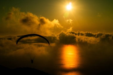 Bir paraglider, sakin bir okyanusun üzerindeki gökyüzünde zarif bir şekilde gezinir, altın günbatımı ışığı, tüylü bulutları aydınlatır ve huzurlu bir atmosfer yaratır..