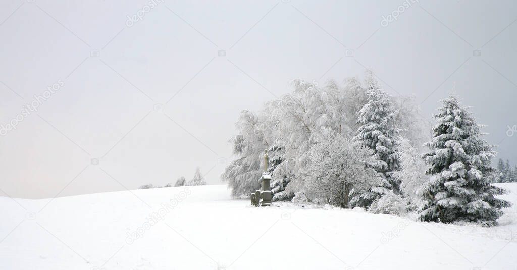 Meadow covered with snow with a line of trees and a roadside cross. Religious thanksgiving sculpture in the mountains.