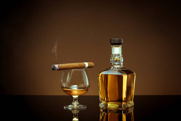 close up view of cigar, bottle of congnac and a glass aside on color back. 