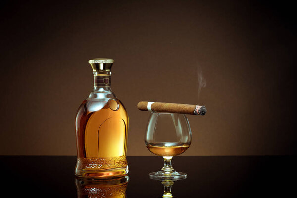 close up view of cigar, bottle of congnac and a glass aside on color back. 