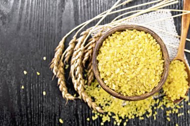 Bulgur Groats - buğday taneleri - kilden bir kase ve kaşık içinde çuval bezi peçetesinde, tahtadan arka planda buğday kulakları