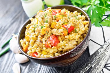 Kıymalı arpa lapası, sarı ve kırmızı dolma biber, sarımsak ve soğan kilden bir kasede, peçete ve maydanozla siyah tahta arka planda.