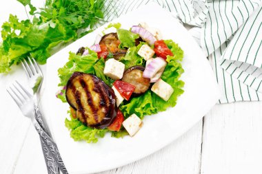 Kızarmış patlıcan salatası, kırmızı soğanlı taze domates ve tuzlu feta peyniri, sebze yağı ve soya sosuyla tatlandırılmış, tabakta marul, peçete, çatal ve beyaz tahta arka planda maydanoz.